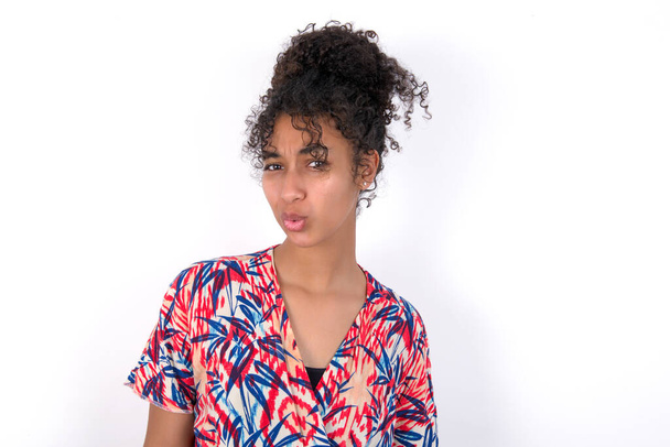 girl expressing disgust, unwillingness, disregard having tensive look frowning face, looking indignant with something. Young beautiful mixed race woman wearing colourful dress over white wall - Photo, Image