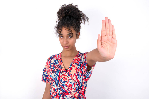girl doing stop gesture with palm of the hand. Warning expression with negative and serious gesture on the face. Young beautiful mixed race woman wearing colourful dress over white wall - Photo, Image