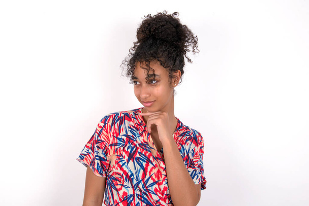 girl looking dreamily and supporting her chin with her pointing finger. Young beautiful mixed race woman wearing colourful dress over white wall - Photo, Image