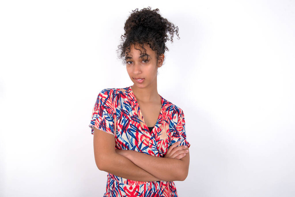 girl biting her lip and crossing her arms. Young beautiful mixed race woman wearing colourful dress over white wall - Photo, Image