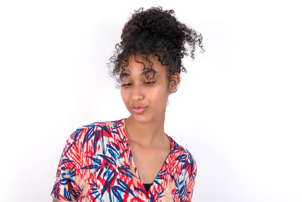 girl looking at the tip of her nose and putting her lips together. Young beautiful mixed race woman wearing colourful dress over white wall - Photo, Image