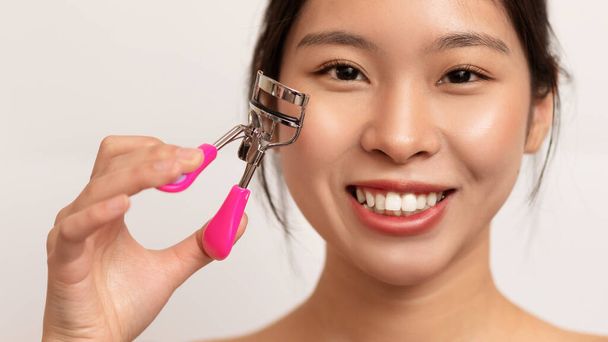 Happy asian lady using eyelash curler, making daily beauty routine, applying eye makeup over white studio background, panorama. Domestic self-care concept - Foto, immagini