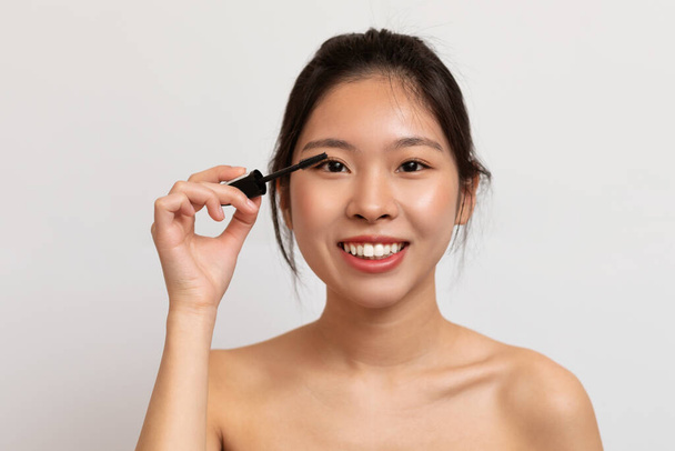 Szczęśliwa koreańska pani stosująca makijaż przy użyciu tuszu do rzęs, ciesząca się rutyną porannego piękna, pozująca na białym tle studia. Kosmetyki do makijażu koncepcja - Zdjęcie, obraz