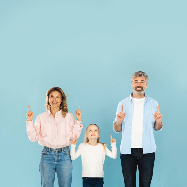 Radostná rodina tří ukazujících prstů nahoru usmívající se na kameru pózující nad modrým pozadím ve studiu. Rodiče a dcera ukazují kopírovací prostor nad hlavou. Čtvercový snímek - Fotografie, Obrázek