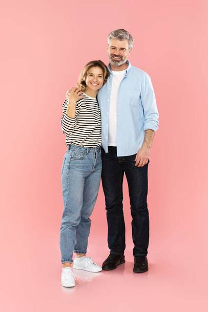 Rakastava keski-ikäinen pariskunta syleilee pysyvän hymyillen kameralle studiossa vaaleanpunaisella taustalla. Aviomies ja vaimo halailevat positiivisia tunteita, pukeutuvat rentoihin vaatteisiin. Rakkaus ja suhde - Valokuva, kuva