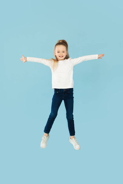 Happy Little Girl Posing In Mid Air Jumping And Spreading Arms Over Blue Background. Vertical Shot, Full Length. Carefree Child Having Fun In Studio. Kids Fashion And Style - Photo, Image