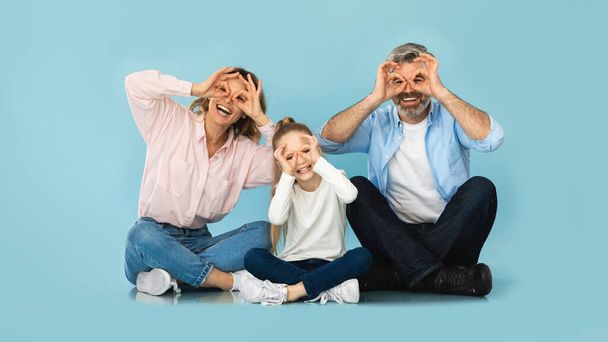 Positive Family Of Three Having Fun Looking At Camera Through Finger Glasses Sitting On Blue Studio Background. Middle Aged Parents And Little Daughter Posing Smiling To Camera. Panorama - Foto, afbeelding