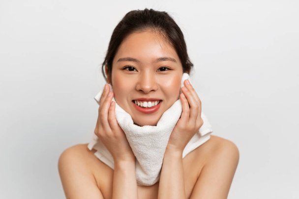 Hermosa mujer asiática disfrutando de la rutina de belleza matutina después de la ducha, tocando su cara con una toalla y sonriendo a la cámara, posando sobre fondo blanco - Foto, Imagen