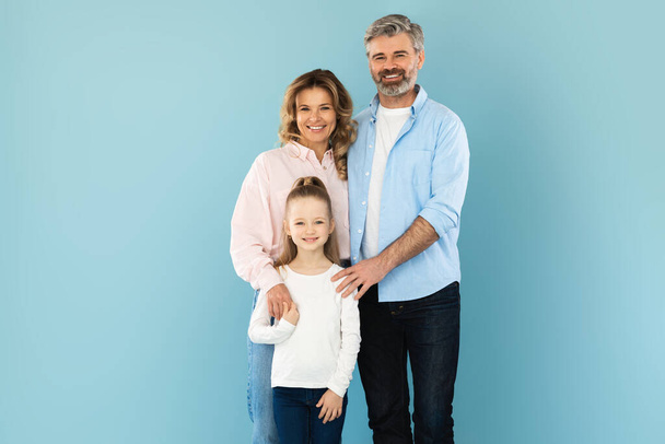 Fröhliche Eltern und kleine Tochter umarmen lächelnd die Kamera und posieren gemeinsam im Studio vor blauem Hintergrund. Aufnahme einer dreiköpfigen Familie, die positive Gefühle ausdrückt - Foto, Bild