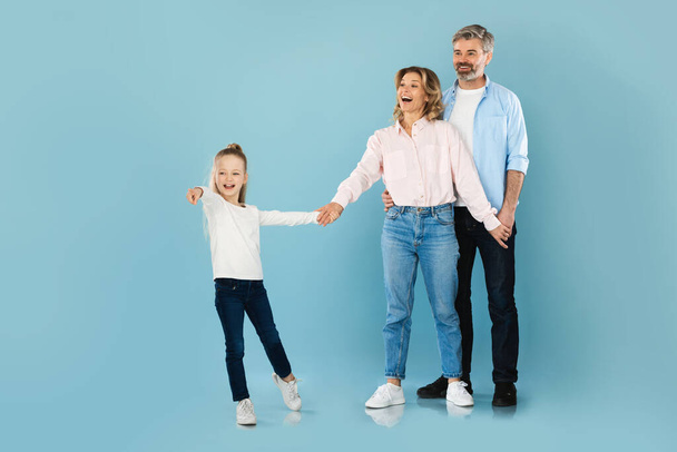 Famille joyeuse avec une petite fille tenant la main et pointant du doigt debout sur fond bleu studio. Les parents et l'offre de publicité pour enfants. Prise de vue complète - Photo, image