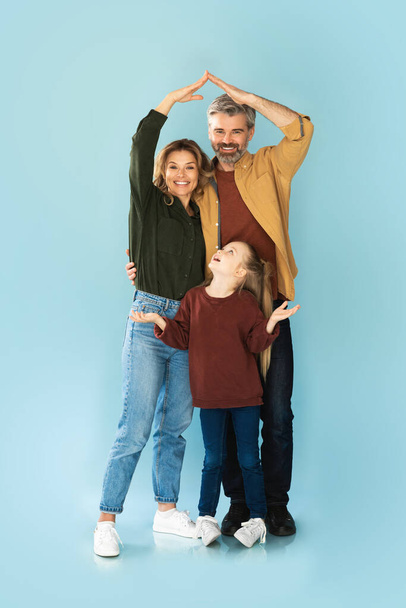 不動産の提供。小さな娘の上に家の屋根を作る腕を作る幸せな両親がスタジオで青の背景の上に一緒に立って.ファミリー・ハウジング・コンセプト - 写真・画像