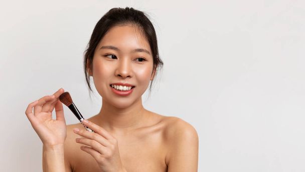 Das tägliche Make-up-Konzept. Porträt einer jungen asiatischen Dame mit natürlicher Schönheit, die ihren Pinsel hält, posiert auf weißem Hintergrund, Panorama mit freiem Raum - Foto, Bild