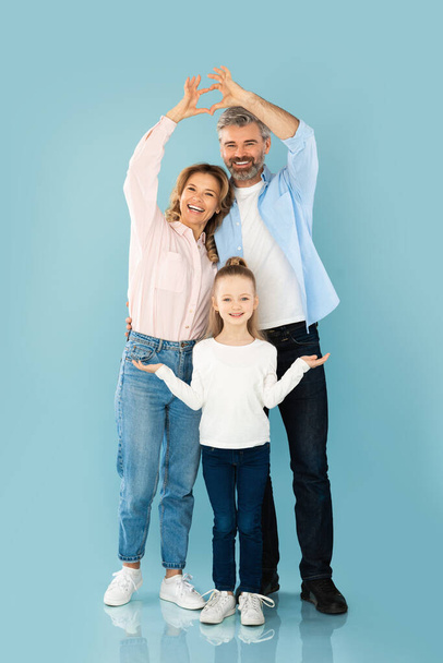Ευτυχισμένοι γονείς ενώνει τα χέρια κάνοντας τα δάχτυλα καρδιά πάνω από τις κόρες τους κεφάλι παριστάνοντας χαμογελώντας στην κάμερα στέκεται σε μπλε φόντο στούντιο. Οικογένεια και αγάπη έννοια - Φωτογραφία, εικόνα