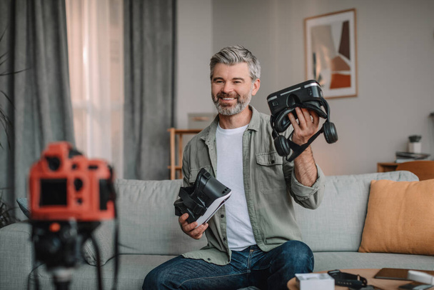 Smiling senior european male with beard shooting blog review on vr glasses in living room interior. Modern work remotely and technology at home, game and entertainment during covid-19 quarantine - Foto, Imagen