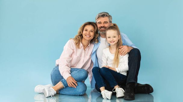 Eltern und kleine Tochter sitzen zusammen und umarmen posieren auf blauem Studiohintergrund. Vater und Mutter im mittleren Alter umarmen ihr Kind lächelnd in die Kamera. Panorama - Foto, Bild