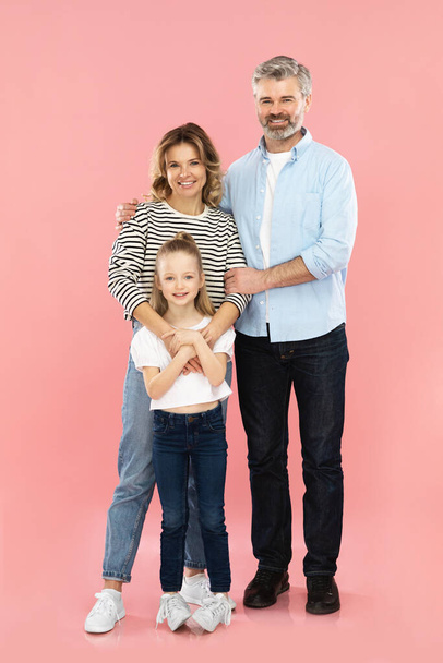 Veselá rodina středověkých rodičů a malá dcera objímající úsměv na kameru stojící nad růžové studio pozadí. Šťastná rodičovská koncepce. Svislý výstřel, plná délka - Fotografie, Obrázek