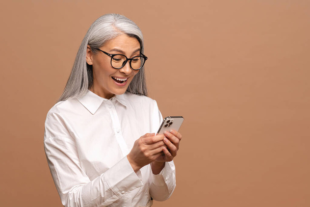 Surprised attractive senior woman holding smartphone, chatting in social networks, texting messages, smiling, reading good news, isolated on beige - Fotografie, Obrázek