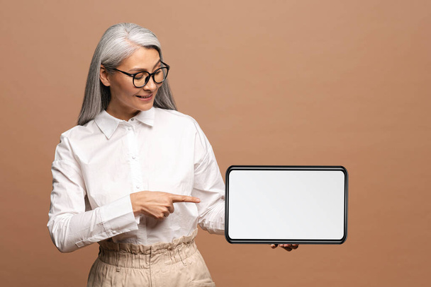 Portrait of the confident senior woman holding tablet and pointing at the screen with smile. Indoor studio shot isolated on beige background - Foto, Imagen