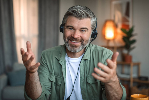 Cheerful mature european male teacher with beard in headset and glasses gesticulates and looks at camera in room interior. Webinar and meeting, work remote, customer support at home during covid-19 - Foto, imagen