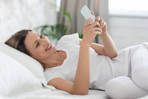 Happy pretty young blonde lady chilling on bed and chatting with her friends, using brand new cell phone and smiling, wearing white pijamas. Communication and modern technologies - Photo, image