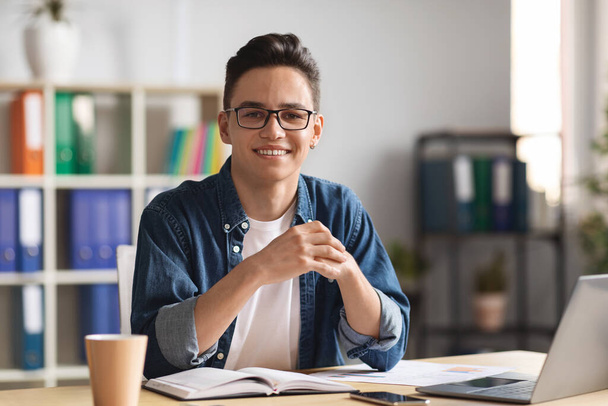 Porträt eines lächelnden jungen männlichen Unternehmers, der mit Laptop am Schreibtisch posiert Im Büroinnenraum sitzt ein hübscher Millennial-Mann mit Brille am Tisch und blickt in die Kamera, Kopierraum - Foto, Bild