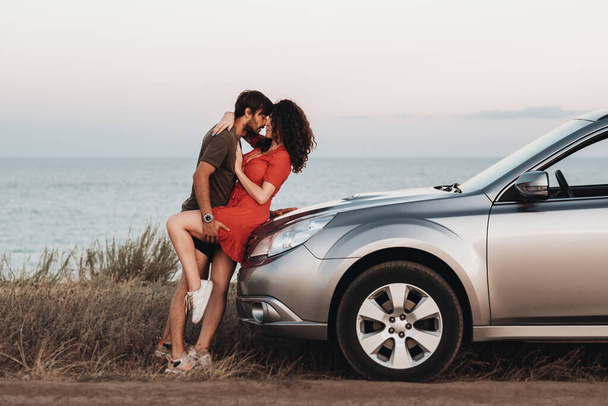 Young Lovers Man and Woman Hugging on the Hood of Their SUV Car on Background of Sea at Sunset - Фото, зображення