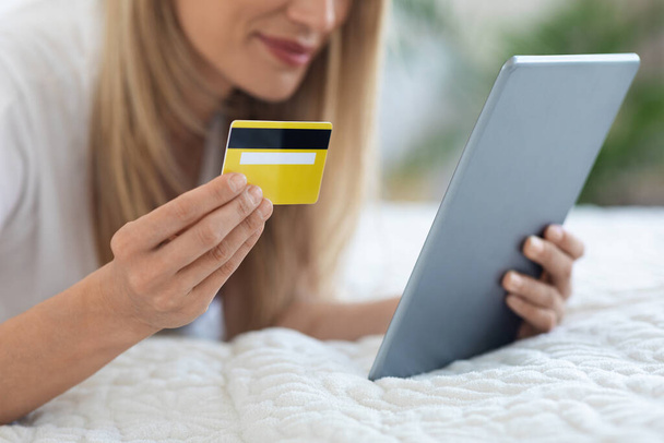 Yeni dijital tablet ve sarı plastik kredi kartıyla yatağa uzanmış, internet üzerinden ürün ve hizmetler için ödeme yapan, kırpılmış, e-ticaret konsepti olan tanınmamış sarışın kadın. - Fotoğraf, Görsel
