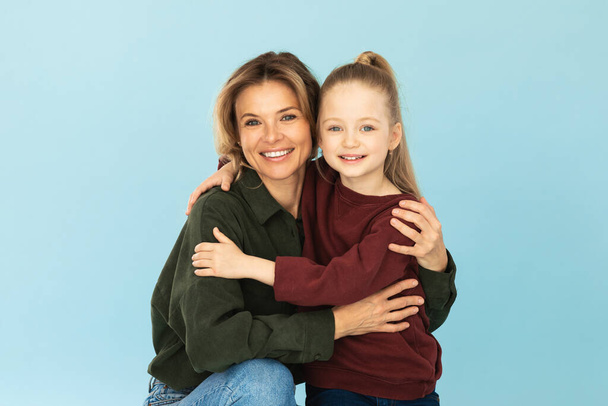 Cheerful Mommy Embracing Adorable Little Daughter Posing Together Standing Over Blue Background. Studio Shot. Mom And Kid Girl Hugging Wearing Casual Clothes Smiling To Camera. Joy Of Motherhood - Photo, Image
