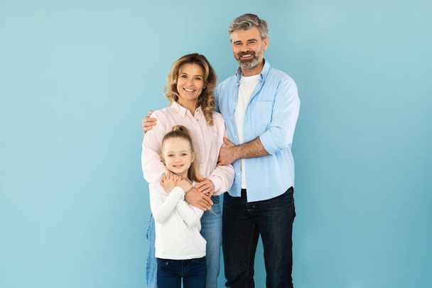 Family Of Three Embracing Smiling To Camera In Studio Standing On Blue Background. Shot Of Happy Middle Aged Parents And Their Little Daughter Posing Together. Happiness Concept - Photo, Image