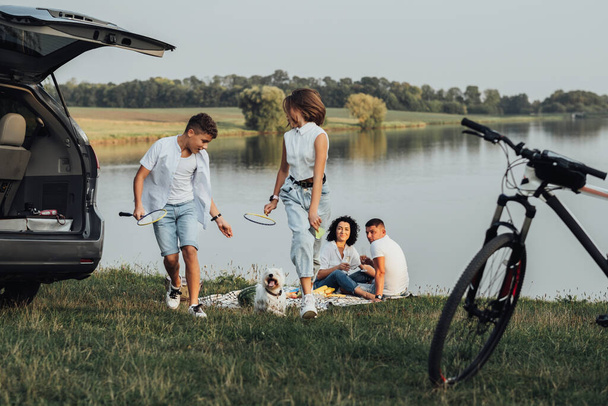 Kaksi teini-ikäistä lasta, poika ja tytär leikkivät lemmikkikoiran kanssa, kun vanhemmat ottavat piknikin järven rannalla, neljä jäsentä onnellinen perhe nauttii viikonlopusta road trip autolla - Valokuva, kuva