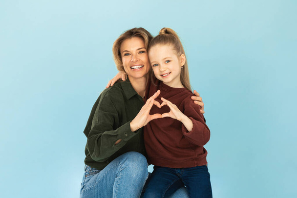 Šťastná maminka a malá dcera spojující prsty, aby se tvar srdce s úsměvem na kameru popěvující objímání na modrém pozadí ve studiu. Rodina a láska, koncepce dne matek - Fotografie, Obrázek