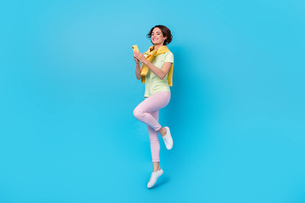 Full size photo of cute millennial brunette lady jump hold telephone wear t-shirt pants footwear isolated on blue background. - Valokuva, kuva