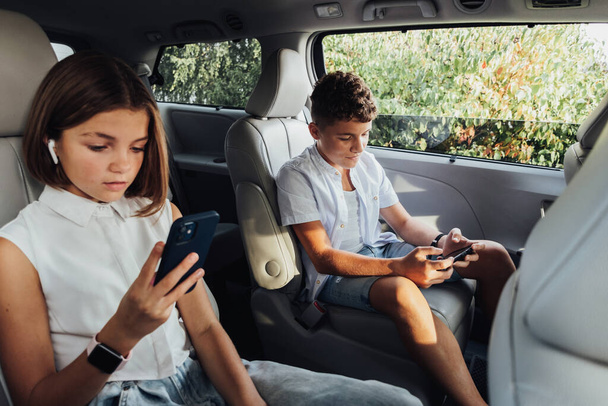 Joven chico caucásico y su hermana adolescente se centró en los teléfonos inteligentes mientras estaba sentado en la minivan, los niños jugando juegos en fin de semana Viaje familiar por carretera en coche - Foto, imagen
