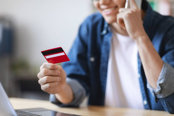 Mobile Payments. Smiling Young Male Holding Credit Card And Taling On Cellphone While Sitting At Desk In Office, Unrecognizable Millennial Man Enjoying E-Commerce And Easy Shopping, Cropped - Photo, Image