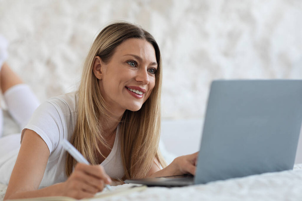 Fröhliche attraktive blonde junge Frau, die von zu Hause aus an einem Online-Webinar teilnimmt, auf dem Bett liegt, Laptop benutzt und Notizen macht, Schlafzimmereinrichtung, Kopierraum. E-Education-Konzept - Foto, Bild