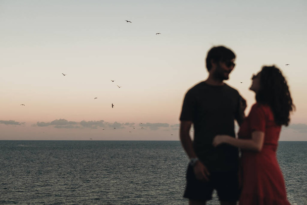 夕方の日没後に海の背景に立つ焦点を当てていない青いカップル、水上を飛ぶカモメ - 写真・画像
