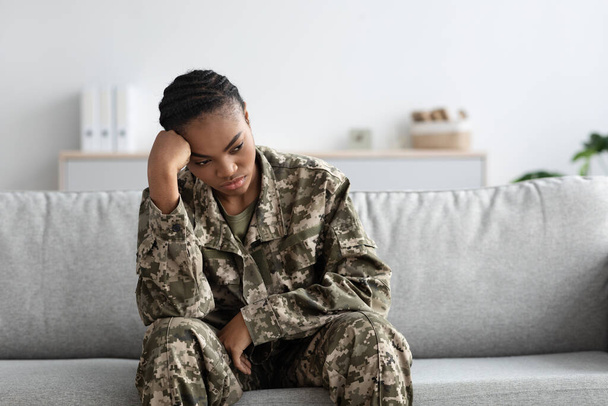 Conceito de depressão. Retrato de soldado feminino preto perturbado sentado no sofá dentro de casa, mulher militar afro-americana deprimida em transtorno de estresse traumático pós sofrimento uniforme, espaço de cópia - Foto, Imagem