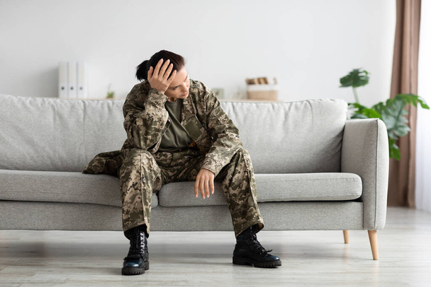 Duševní poruchy ve vojsku. Depressed voják dáma v uniformě sedí na gauči Indoors, Portrét Pensive rozrušená armáda žena nosí maskování dotýkání hlavy a dívá se pryč, kopírovat prostor - Fotografie, Obrázek