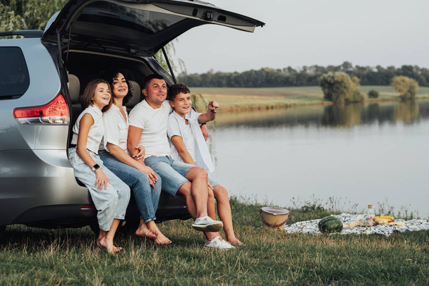 Happy Four Caucasian Members Rodzina siedzi w bagażniku minivana samochodu, matka i ojciec z dwójką nastoletnich dzieci, syn i córka o weekend piknik na świeżym powietrzu nad jeziorem - Zdjęcie, obraz