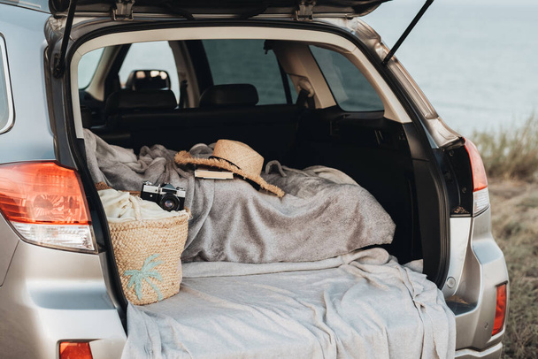 Plaid mit Hut und Kamera und Strandtasche im geöffneten Kofferraum des SUV-Autos, das an der Küste parkte, Sea Weekend Concept - Foto, Bild