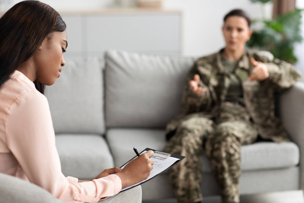 Μαύρη γυναίκα ψυχοθεραπεύτρια σημειώνει κατά τη διάρκεια της συνεδρίας θεραπείας με τη στρατιωτική γυναίκα, στρατιώτης κυρία με στολή κάθεται στον καναπέ και μοιράζονται τα προβλήματά της με ψυχίατρο κατά τη συνεδρίαση στο γραφείο - Φωτογραφία, εικόνα