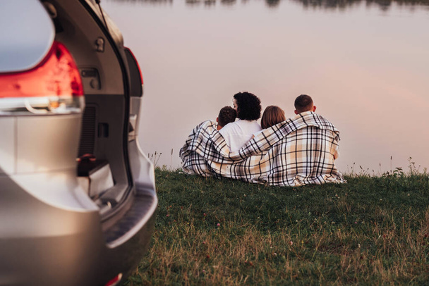 夕日の湖で草の上に座っている4人の家族、週末のロードトリップを楽しむ2人の子供と母と父 - 写真・画像