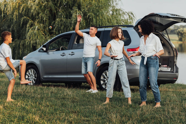 Feliz familia de cuatro miembros que se divierten al aire libre, madre bailando con la hija adolescente mientras el padre juega con el hijo, coche minivan en el fondo - Foto, Imagen
