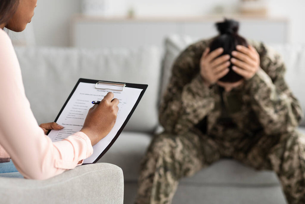 Black Psychiatrist Lady Filling Questionnaire During Therapy Meeting With Depressed Military Woman, Soldier Female With Mental Problems Sitting On Couch And Touching Head In Despair, Selective Focus - Photo, Image