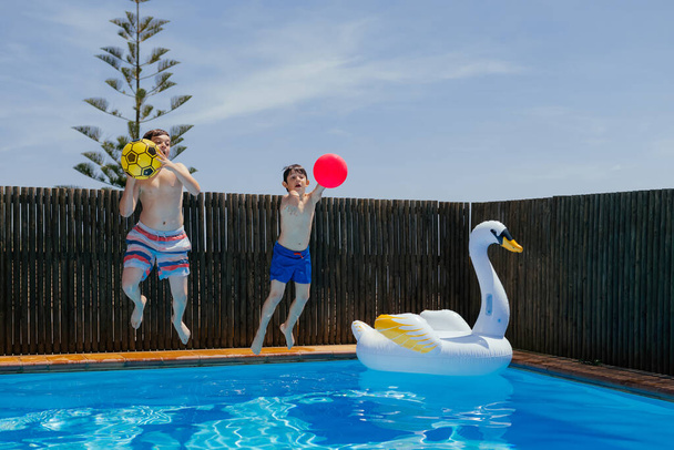 Children play with a balls and jump into a blue water pool, on a sunny summer day during holidays. Children playing water sport games on home backyard with inflatable swan. Horizontal. - Fotografie, Obrázek