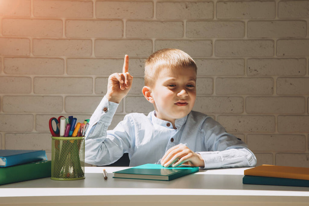 Egy fehér asztalnál ülő iskolás fiú kézmozdulatot tesz, hogy van egy ötlete. Iskolai koncepció - Fotó, kép