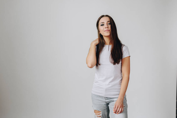 Young Woman standing on the Right side in front of a white background in jeans and a basic t-shirt. Girl with makeup cute straightens her hair. Hair care concept. - Photo, Image
