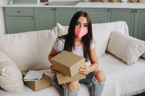 写真上から、ピンクのマスクの女の子は手に箱でソファに座っています。自宅で検疫作業中の少女。枕付きソフトライトソファ. - 写真・画像