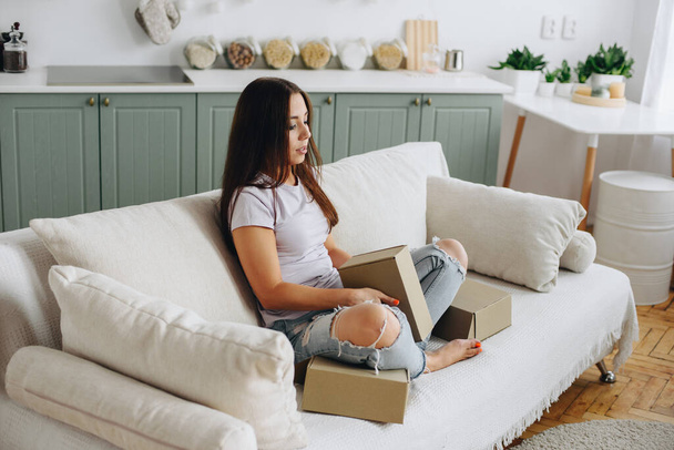young woman in torn jeans sits pensively on sofa. girl is holding boxes in her hands. Concept Unemployed. Home cozy interior. - Фото, зображення