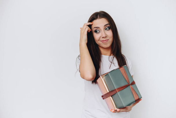Brunette Woman Holds Gifts in Her Hands. Think about what to give for Holiday. Girl Scratches her Forehead and Grimaces. Girl on White Background. - Photo, Image